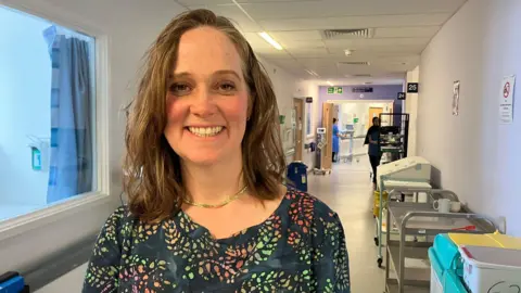 A woman wearing a flowery top and gold chain with a hospital ward behind her. She has shoulder length brown hair and is smiling at the camera. 