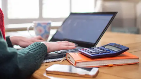 PA Media A woman wearing a green wool jumper and a red scarf sits at a table and works on a laptop. She has a coffee mug with flowers on it next to her. An orange diary, blue calculator and white mobile phone are also on the wooden table.