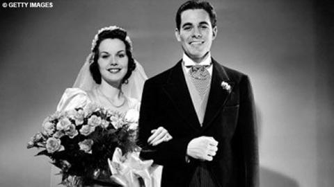 A bride, holding a bouquet, and groom stand with their arms linked