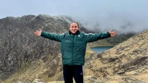 Elissa Hubbard successful  a greenish  sports overgarment   and achromatic  Adidas tracksuit bottoms stands astatine  the apical  of a upland  and a water  is besides  disposable   successful  the background. She has her arms outstretched and is smiling 