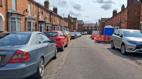 LDRS Cars parked on both sides of a residential street. There is a skip and and portaloo with a barrier around it in between some of the cars. It is a bright day with some clouds.