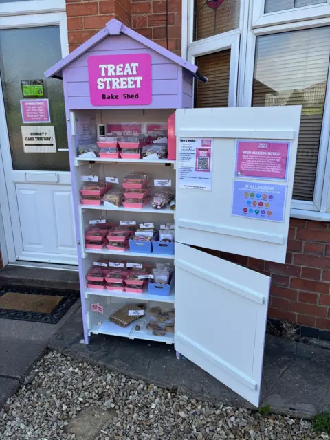 The Treat Street stall. It is a tall and thin shed, lilac in colour and consists of six shelves on which treats including cakes, cookies, blondies are stored. The two doors are swung open.
