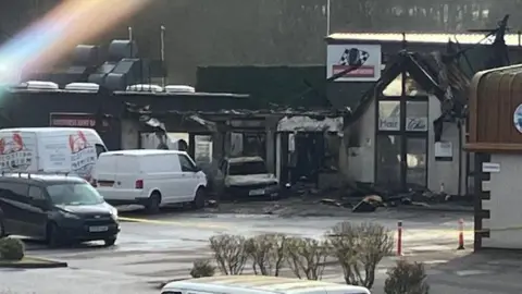 A photo of the burnt out shops and car taken from behind a fence