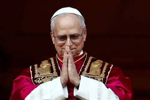 Pope Leo XIV prays on the balcony of St Peter's He is wearing a white cap and crimson cape 