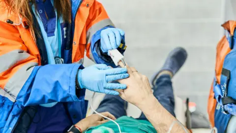 A female paramedic wearing a orange and blue coat and blue shirt and trousers is reading the heartbeat of a patient lying down on a stretcher with an oximeter - a white device on their finger they are holding up.