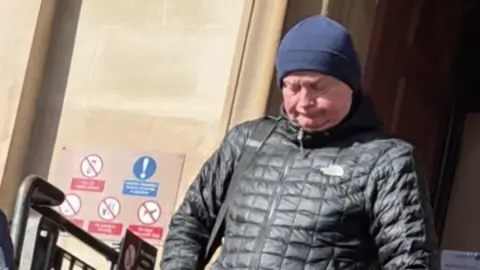 A man of middle years in a blue wool hat and black puffer-style jacket. He is leaving a blonde sandstone building with his head bowed as though he does not want to be photographed.