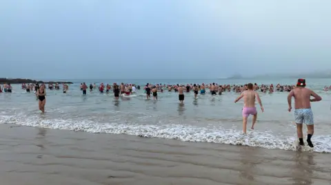 About a hundred swimmers getting into the sea, some wearing christmas hats. The beach is foggy.