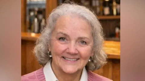 Julie has short curly grey hair and is wearing a pink jumper with squares on it and a while collared shirt underneath. She is smiling at the camera and there is a wooden bar behind her
