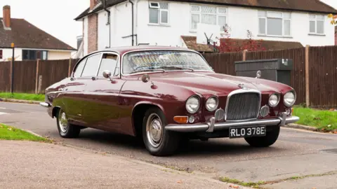 A maroon Jaguar car parked on tarmac with a wooden fence and small patch of grass behind it. 