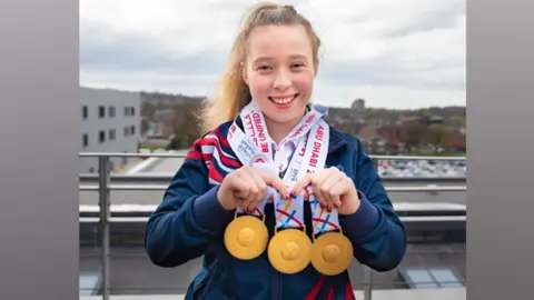 Kiera Byland Kiera Byland is smiling directly at the camera. Around her neck are three gold Special Olympics medals which she is holding up towards the camera.