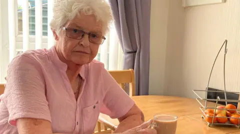 Sandra Robinson is wearing a pink shirt and is holding the handle of a mug which has a brown drink in it. On her dining room table is a fruit stand which has oranges inside.