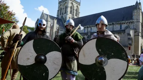 Grim Falfest 2023 Three men dressed as Vikings, with steel helmets, black-and-white round shields, axes and spears, in front of St James' Church in Grimsby.