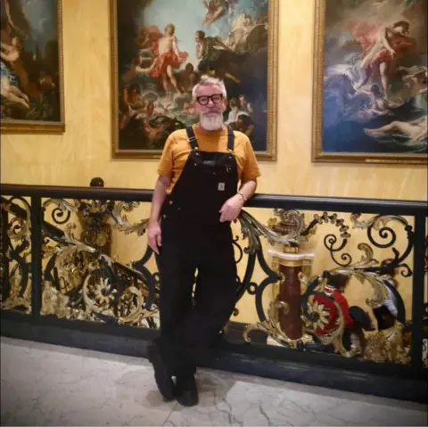 Mary Courtney Simon Gee wears a mustard yellow t-shirt with black dungarees. He has thick set glasses and a grey beard and mousache