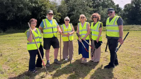 A group of volunteers stood in the park holding their garden tools and wearing high vis jackets.