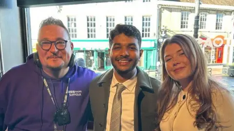 Wayne Bavin, wearing headphones, glasses and a purple BBC Radio Suffolk jumper, standing next to Gouse Subhan Saheb, who is wearing a green jacket over a shirt and tie, and Katie Molloy, who is wearing a white dress. They are all smiling and looking into the camera.