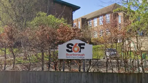 A sign for Scarborough Sixth Form College is displayed in front of a wooden fence and leafless shrubs. The sign features a bold ‘S6F’ logo with an orange and red circular design, and the text ‘Scarborough Sixth Form College’ underneath.