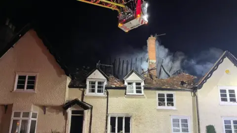 Suffolk Fire and Rescue Service A large yellow property at night time. A firefighter stands on an aerial ladder above the property to tackle a blaze in the home. Smoke can be seen billowing out from the roof. 