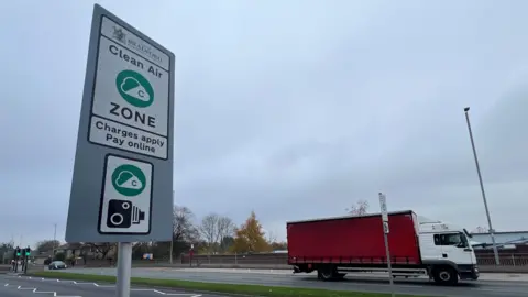 A road sign indicates Bradford's Clean Air Zone. It is captioned "Charges apply, pay online". A red lorry drives along the dual carriageway in the background.