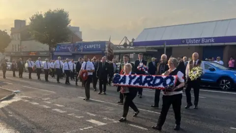 Two women carry a wreath with the words Bayardo on it in blue on a white background. A man is behind them carrying a yellow floral tribute. There are people behind them dressed in black and white.