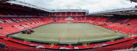 Simon Hutton Old Trafford stadium, the famous red seats and stands incase the pitch. On the pitch is two tractors which are laying turf.