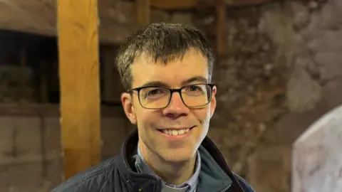 Luke Deal/BBC A head and shoulders image of Reverend Daniel Whiffin. He is wearing glasses and smiling into the camera. He appears to be standing inside the tower of the church.