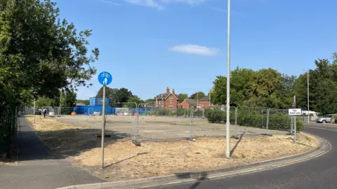 The derelict land where the coffee shop will be built. It has concrete paving surrounded by grass and with metal fencing around the site