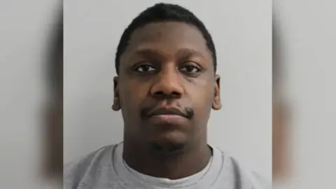 A mugshot style image of a man. He has a blank expression and is staring down the camera. He is wearing a plain grey jumper and has a moustache and short hair