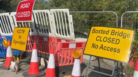 A road has been closed and cordoned off using metal and plastic fending. A row of orange traffic cones has been put in place to further block off access to the road. In front of these cones are metal signs. On the left there are two signs stacked on top of each other. At the bottom is a yellow metal sign with black writing which says, CCTV In Operation. On top of it, is a red metal sign with white writing which says, ROAD CLOSED. On the right of the picture there is a yellow metal sign which says, Boxted Bridge CLOSED No Access.