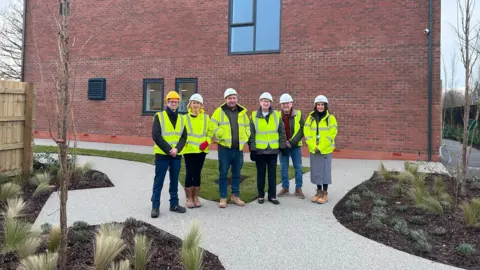 A group of six people are stood on a paved footpath, which is interspersed with several flowerbeds and a lawned area. The people are each wearing fluorescent yellow jackets as well as hard hats. Behind them is a large brick built building.