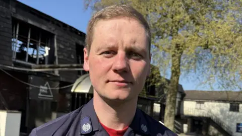 Paul O'Gorman/BBC A young man with sandy blond hair stands wearing a navy uniform. He is standing in front of fire-damaged building. 