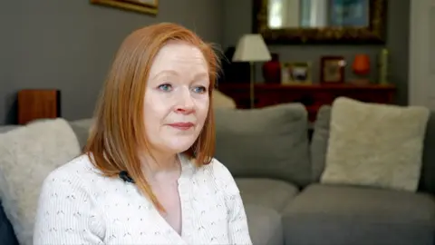 Lorna Pirozzolo, in her late 40s. She has shoulder‑length light brown hair and is sat on a grey sofa in a living room. She's wearing a light-coloured, textured top. Behind her, the room includes a wooden sideboard with framed photos, a lamp, and decorative items. Cushions are arranged on the sofa, and a large framed mirror hangs on the wall in the background.