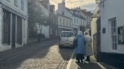 BBC Two elderly people walk away up a cobbled street with a car parked on the rights and buildings either side.