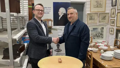 Two men - Niall Fry on the left who is wearing a white checked shirt and a grey blazer, and Marios Theocharous, who is wearing a dark shacket with a dark shirt beneath and has grey hair - shake hands as they stand in an auction house. In front of them, there is a small pine wooden table, with a small jug on it.