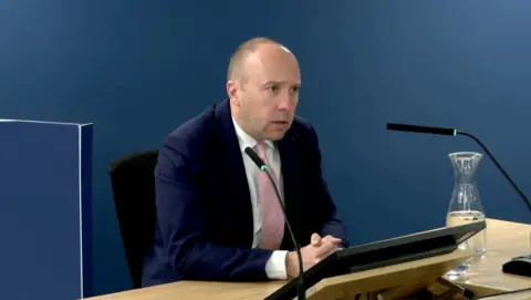 PA Media Matt Hancock, wearing a blue suit and a pink tie with a shaved head, is sitting at a desk at the Covid inquiry while giving evidence.