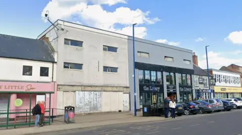 A weathered white building with several small windows and a boarded-up ground floor is joined to a modern-looking grey shopfront with large glass windows and signage, followed by additional businesses. Several cars are parked along the street, while a few pedestrians walk along the pavement. The road has marked lanes and parking areas, and tall blue streetlights line the pavement. Above, the scene is set against a bright blue sky with scattered white clouds.