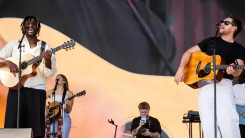 BBC / James Watkins Myles Smith wears a white shirt and black trousers whilst holding a guitar and singing on stage at Radio 1's Big Weekend in Sunderland. Niall Horan joins him, wearing a black t-shirt and white trousers, also holding a guitar and singing. The band for Myles Smith is in the background.