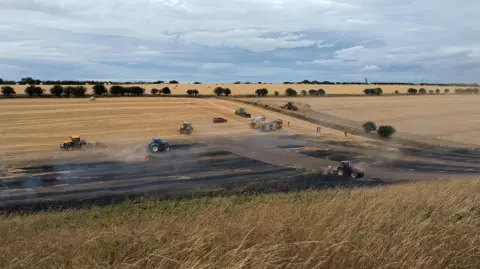Kate Balchin A partially charred field can be seen down a gentle slope, with long grass in the foreground of the photo. Smoke is rising from the blackened ground, and there are six tractors around or on it. There are also two fire engines and a car parked on the field. There is a hedge and another untouched field to the right of the image.