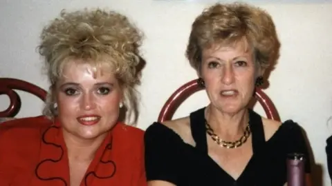 Family handout A printed photo of two women, Tracey and Wendy Speakes. It looks like the late eighties. Tracey is wearing a red blouse with a wavy collar with a black trim. Her hair is white-blond and permed. Wendy has shorted blond hair. She wears sparkly black earrings, a gold chain necklace and off the shoulder black top.