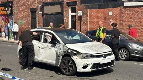 A white Volkswagen Polo which has been badly damaged at the front on the left. The driver's door is also warped. The bonnet is covered in dirt. There is an officer dressed in black leaning into the back window and a police officer in a hi-vis yellow vest looking at the bonnet while standing the other side of the car. There are two other officers standing behind the car. There is a silver car parked on the side of the road to the right and two people walking down the path behind it. There are red brick buildings lining the street with a colourful shop on the corner on the left.