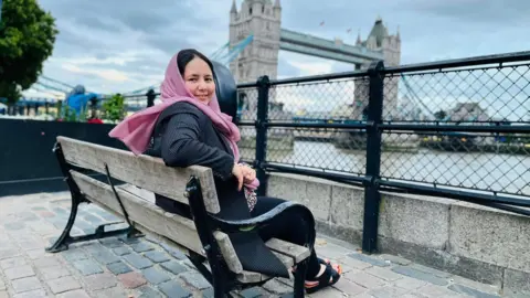 Zahra Joya Zahra sits on a wooden and black metal bench on a cobbled street by the River Thames. Tower Bridge is in the background. She turns and smiles at the camera. She wears a pink headscarf and a black pinstriped coat, and black sandals.