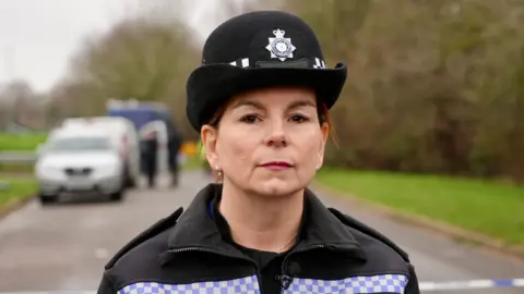 Ant Saddington/BBC Ch Insp Kim Jackson wears a black police force hat with badge and a black police jacket with blue and white checks across the chest. She stands in front of police tape preventing entry to the crime scene in the park.
