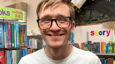 Close-up portrait of a person wearing black-framed glasses and a white T-shirt, standing in a library. Bookshelves filled with colourful books are visible behind them, along with signage reading “authors R–Z” and “Story.” The image is well lit and focused on the person, with library displays and children's reading materials in the background.