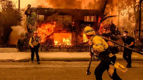 Getty Images Seorang petugas pemadam kebakaran menyeret selang menuju rumah yang terbakar di daerah Altadena di Los Angeles, sementara petugas pemadam kebakaran lain melihat