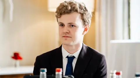 Sam Carling looks slightly off camera, he wears a black suit with a dark tie and white shirt. A soft yellow light is behind him. 