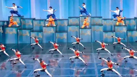 CCTV A screengrab shows robots in gold and red vests performing martial arts on stage during a Lunar New Year showcase, alongside young athletes carrying out a similar routine in the background.