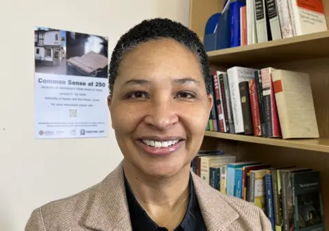 A woman in a brown jacket and dark blouse smiles at the camera in front of a bookshelf