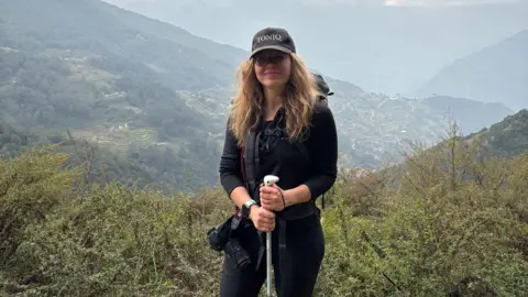 A woman is standing on a mountain holding a walking stick. She is surrounded by green plants and hills in the background. 