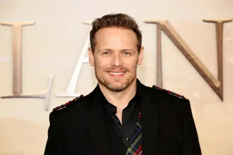 Getty Images A man is smiling at the camera. He's wearing a black blazer and shirt it was tartan elements added to it