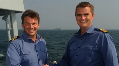 Tom Sharpe Two men in blue Royal Navy uniforms looking at the camera on board a ship with the ocean and a second ship in the background 