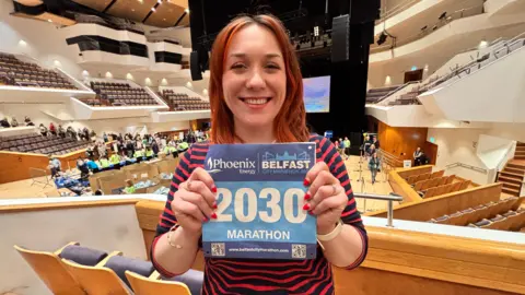 A red haired woman stands inside a large auditorium holding a Belfast City Marathon 2026 race bib showing the number 2030 with the word “MARATHON.” She has a navy and red stripped top on. The venue has tiered seating, wooden railings, and a stage with lighting equipment, while event volunteers in green shirts and tables with race materials are visible in the background.
